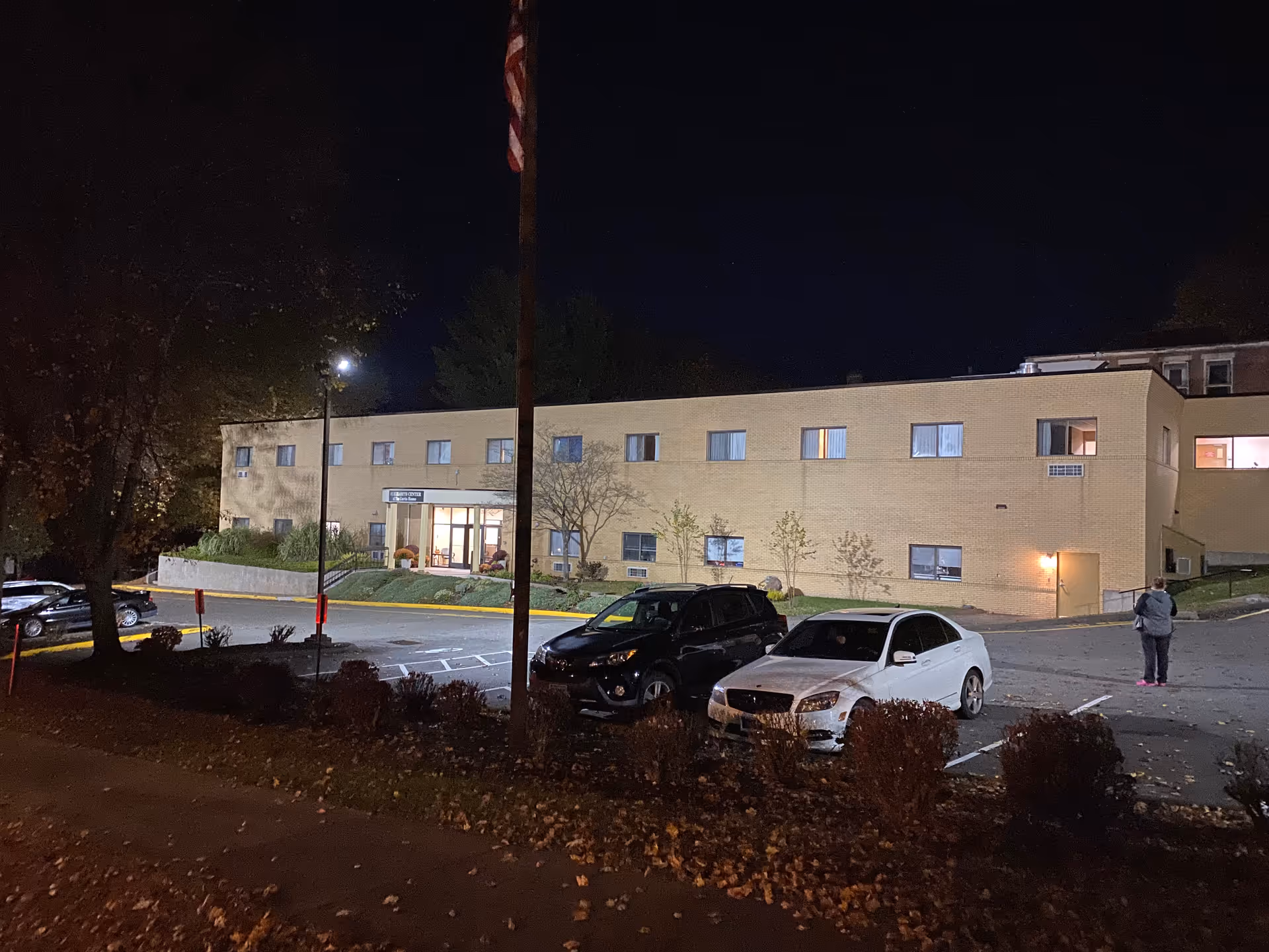 Nighttime exterior view of a two-story beige brick building with several windows lit from inside. There is a parking lot in front with a few parked cars and a person standing near the right side of the image. Trees and bushes surround the area, and an American flag is visible on a pole near the center.