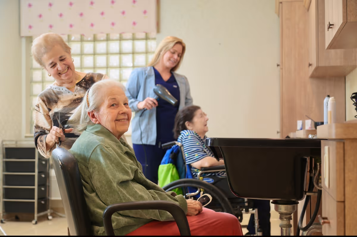 An elderly woman sitting in a salon chair smiling while another woman is cutting her hair. In the background, a caregiver is blow-drying the hair of another elderly woman seated in a wheelchair. The setting appears to be a hair salon area within a senior living facility.
