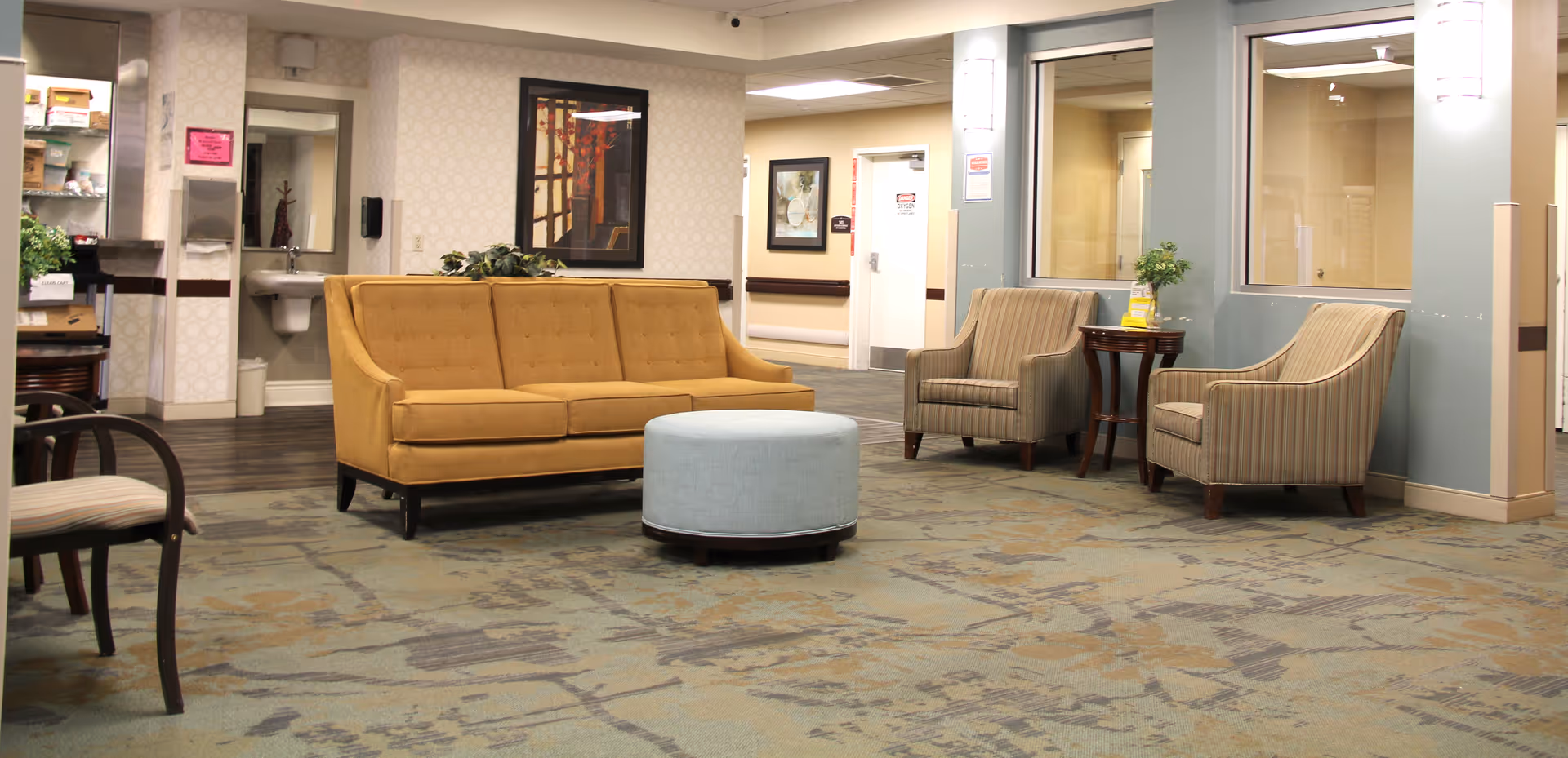 A seating area in a senior living facility with a mustard yellow sofa, two striped armchairs, a round light blue ottoman, and a small wooden side table with a plant. The room has patterned carpet, light-colored walls, framed artwork, and a sink with a mirror in the background.