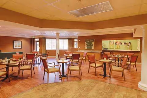 Interior dining room with round tables and red chairs in a senior living facility.