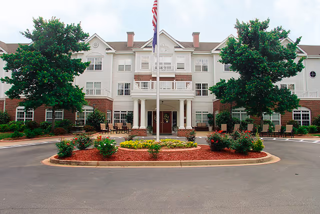 Front exterior view of The Gables senior living facility showing a three-story building with white siding and red brick accents. There is a circular driveway with a landscaped island featuring flowers and shrubs, and two large trees on either side. An American flag is flying on a flagpole in the center of the driveway.