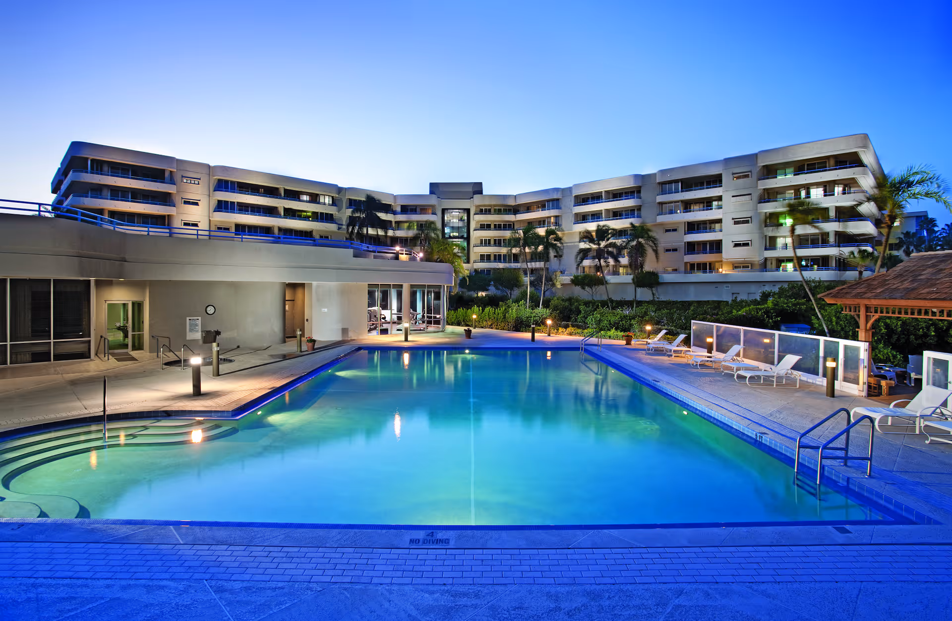 Outdoor swimming pool and deck with lounge chairs in front of a multi-story residential building at dusk.