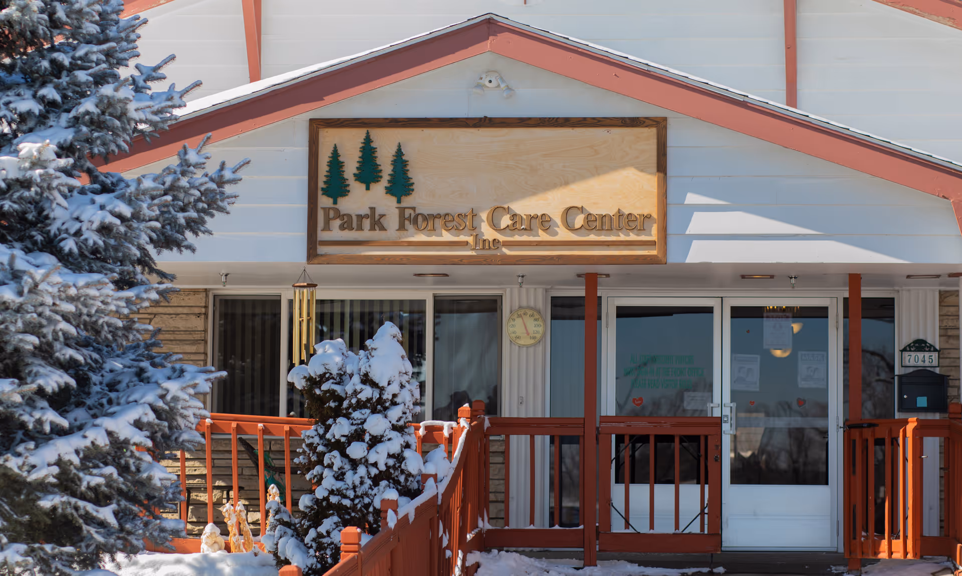 Entrance of Park Forest Care Center, Inc with a wooden sign displaying the facility name above the door. Snow-covered trees and bushes are visible in front of the building, and a red railing lines the walkway leading to the entrance.