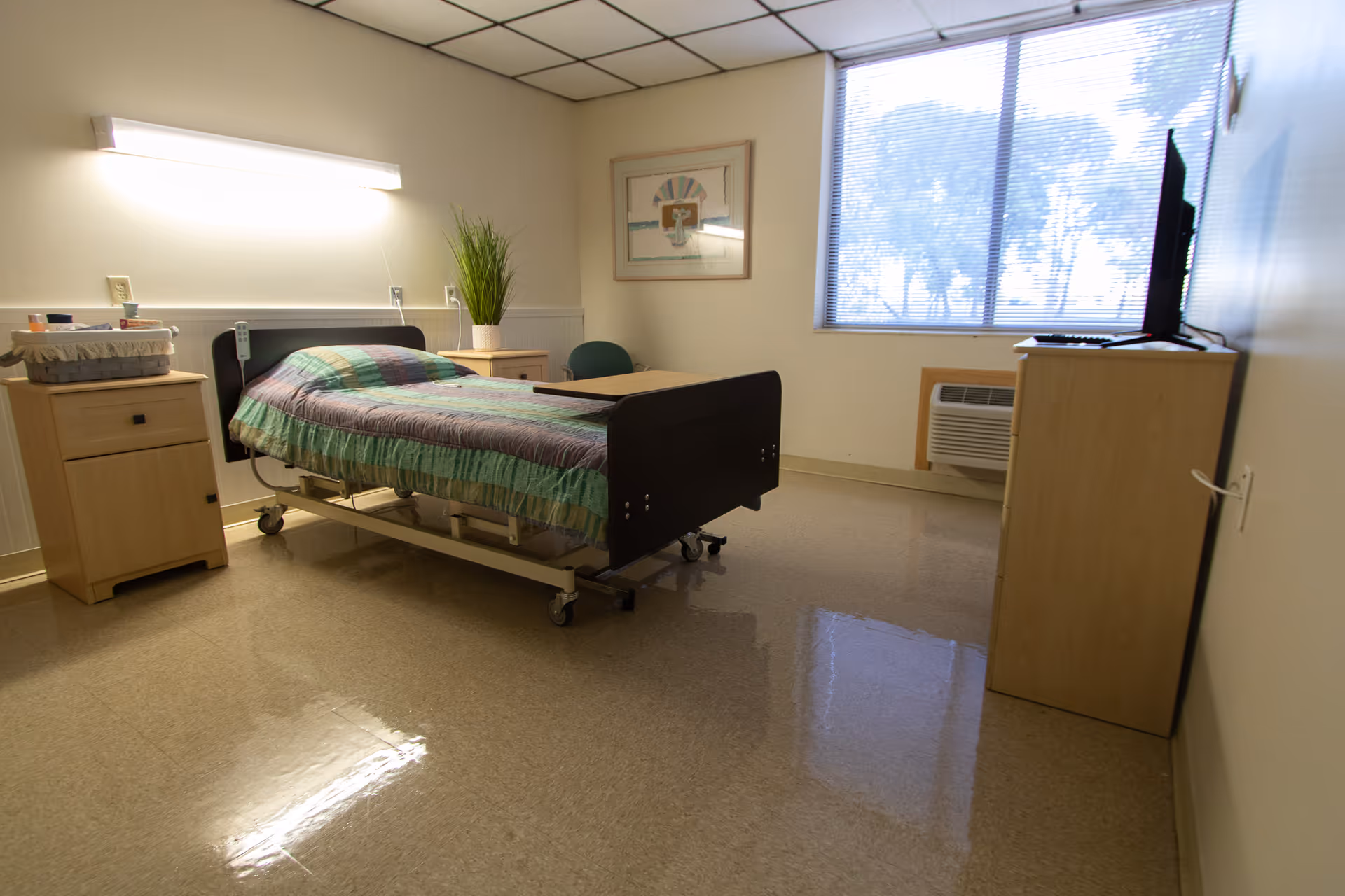 A simple, clean patient room with a hospital bed covered in a green and purple striped blanket. The room has a wooden nightstand with a basket and a potted plant, a small table with a chair, a window with blinds, an air conditioning unit below the window, and a wooden cabinet with a TV on top. The walls are light-colored and there is a framed picture hanging on the wall.
