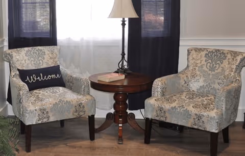 Two patterned armchairs with a floral design are positioned on either side of a round wooden table with a lamp and a book on it. One chair has a dark pillow with the word 'Welcome' embroidered on it. Behind the chairs is a window with sheer white curtains and dark drapes.