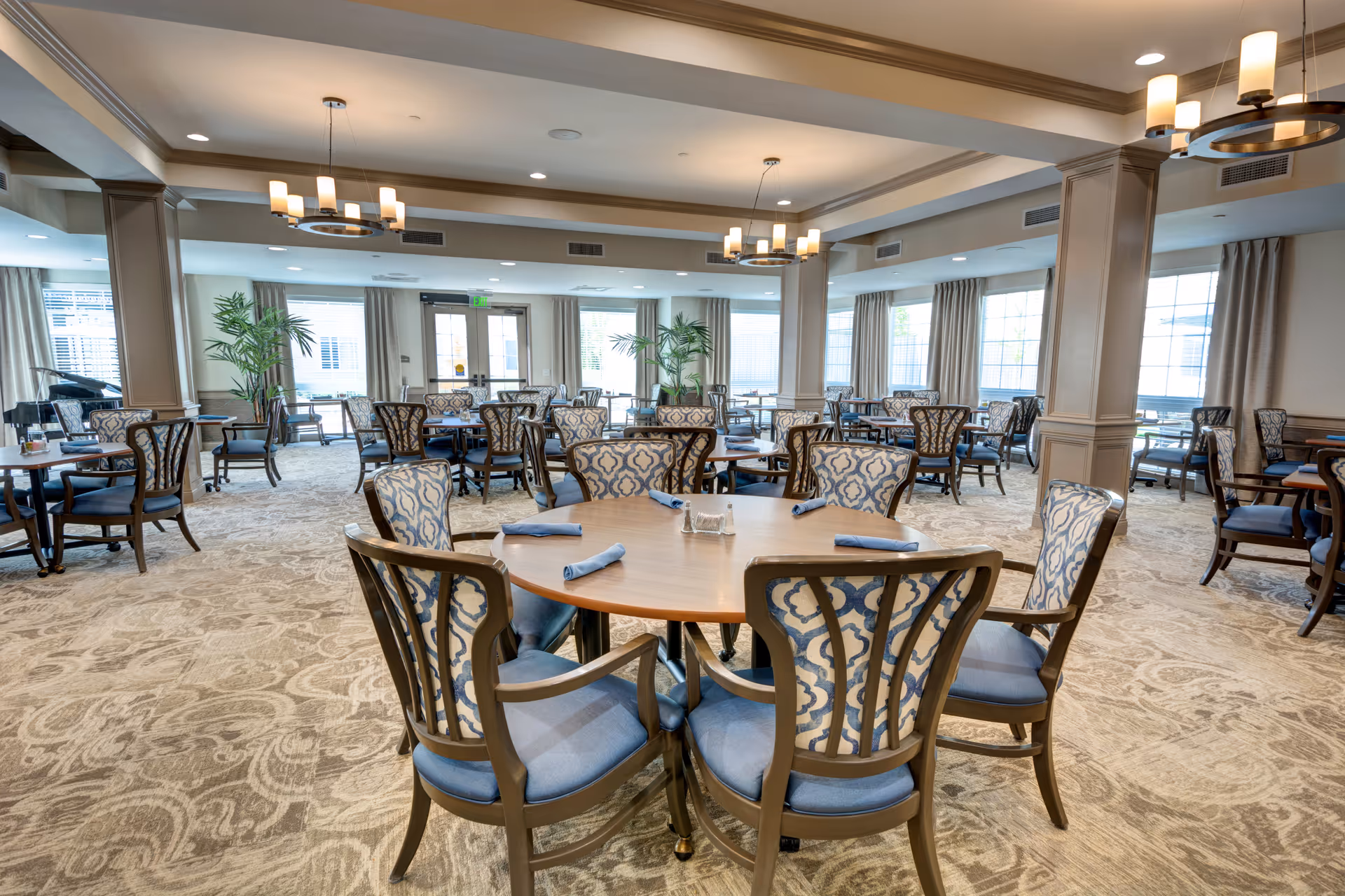 A spacious dining room with multiple round wooden tables surrounded by cushioned chairs with patterned backs. Each table is set with blue cloth napkins and a small centerpiece. The room has large windows with beige curtains, beige carpet with a subtle pattern, and modern ceiling light fixtures. There are also a few potted plants near the windows.