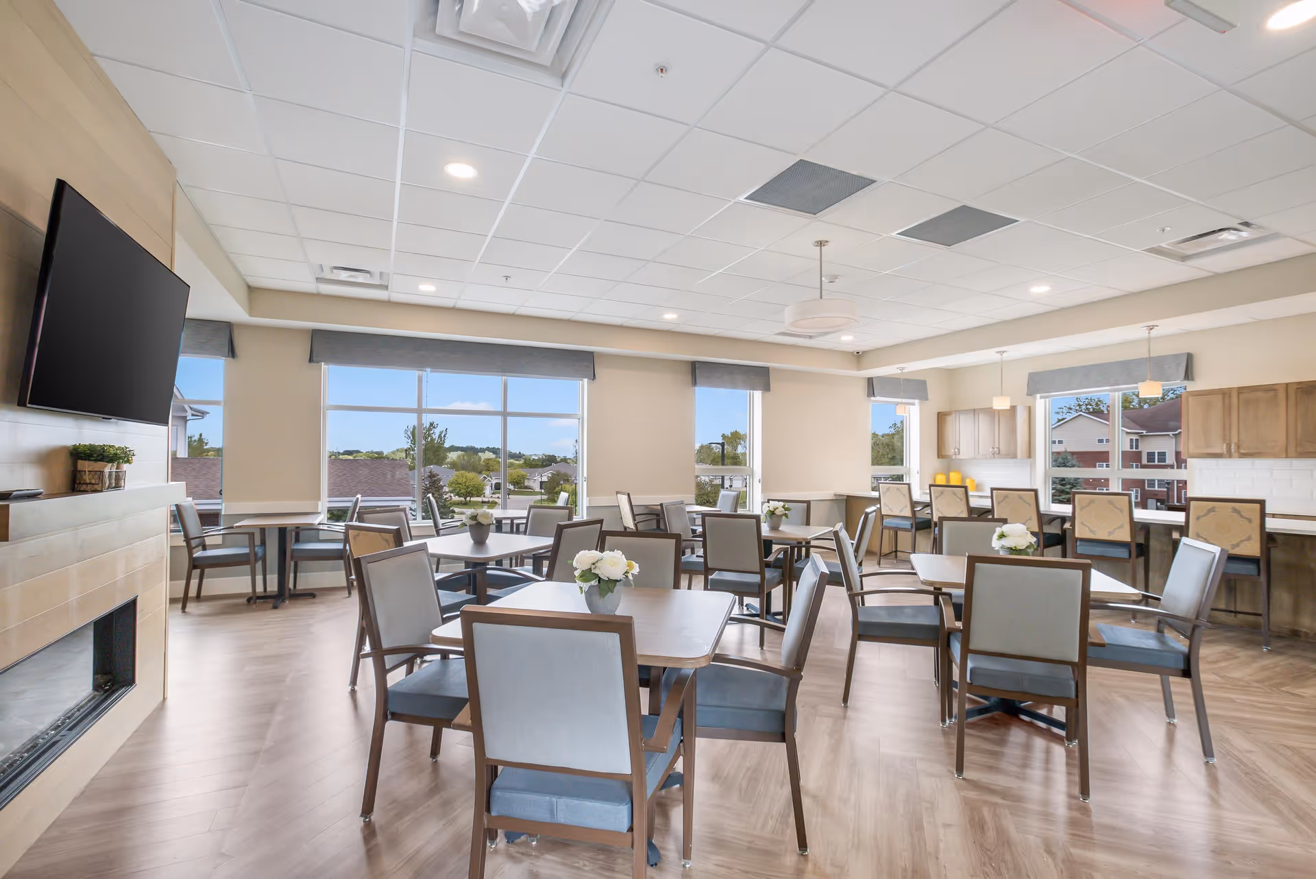 A bright and spacious dining room with multiple square tables and chairs arranged neatly. The room features large windows with views of trees and buildings outside, allowing natural light to fill the space. There is a mounted flat-screen TV above a modern fireplace on one wall, and a kitchen area with a counter and bar stools in the background. The ceiling has recessed lighting and pendant lights over the counter. Small flower arrangements are placed on the tables.