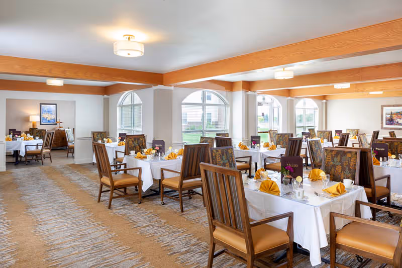 A bright dining room with multiple tables set with white tablecloths, folded yellow napkins, and wooden chairs beneath wood-accented ceiling beams.