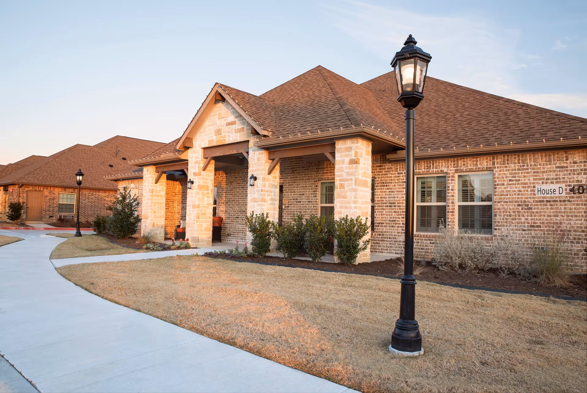 Exterior view of a single-story brick building with a covered porch supported by stone pillars, a black lamp post on a grassy lawn, and a concrete walkway curving in front of the building during daylight.