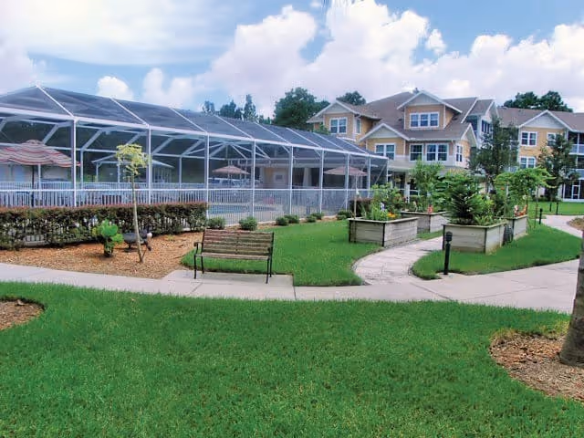 Outdoor area of a senior living facility featuring a well-maintained lawn, a wooden bench, raised garden beds, paved walkways, and a large screened-in pool enclosure. In the background, there are multi-story residential buildings under a partly cloudy sky.