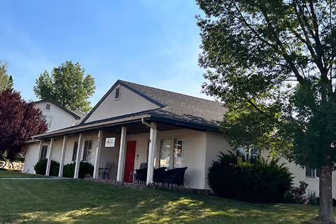 Single-story senior living home with a covered front porch, red door, lawn and trees.