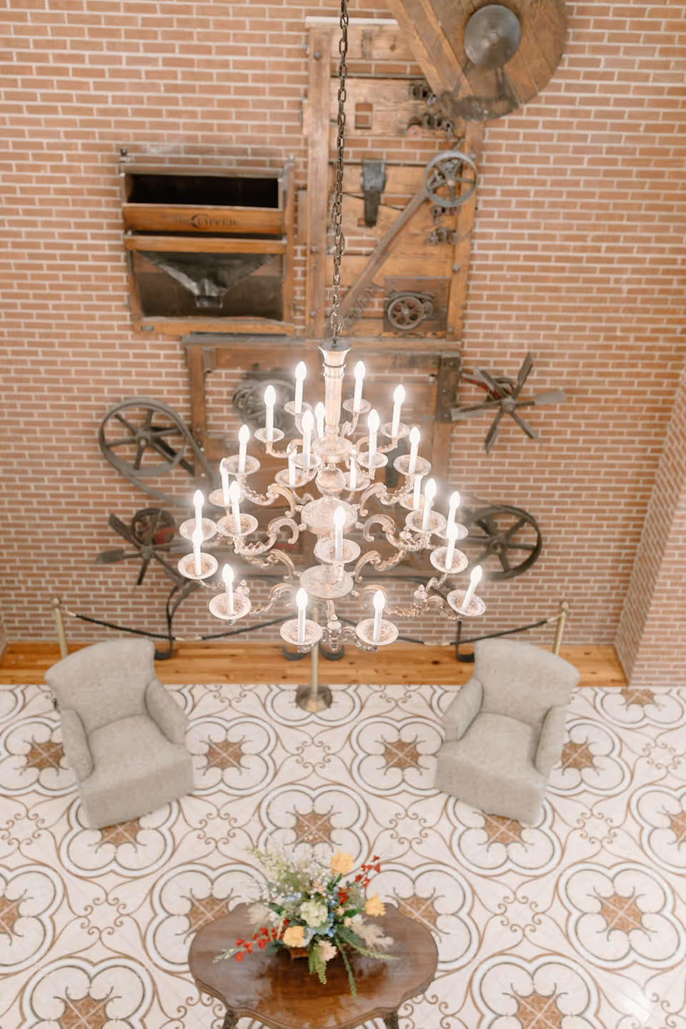 An ornate chandelier hangs above a tiled lobby with two armchairs, a small wooden table with a floral arrangement, and a brick wall decorated with vintage machinery.
