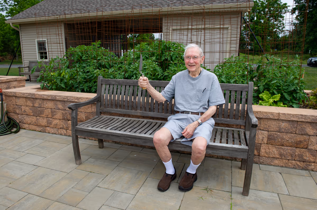 An elderly man wearing a gray t-shirt, gray shorts, white socks, and brown shoes is sitting on a wooden bench outdoors. He is smiling and holding a small gardening trowel in his right hand. Behind him is a raised garden bed with green plants and a trellis structure. A beige building and some outdoor furniture are visible in the background.