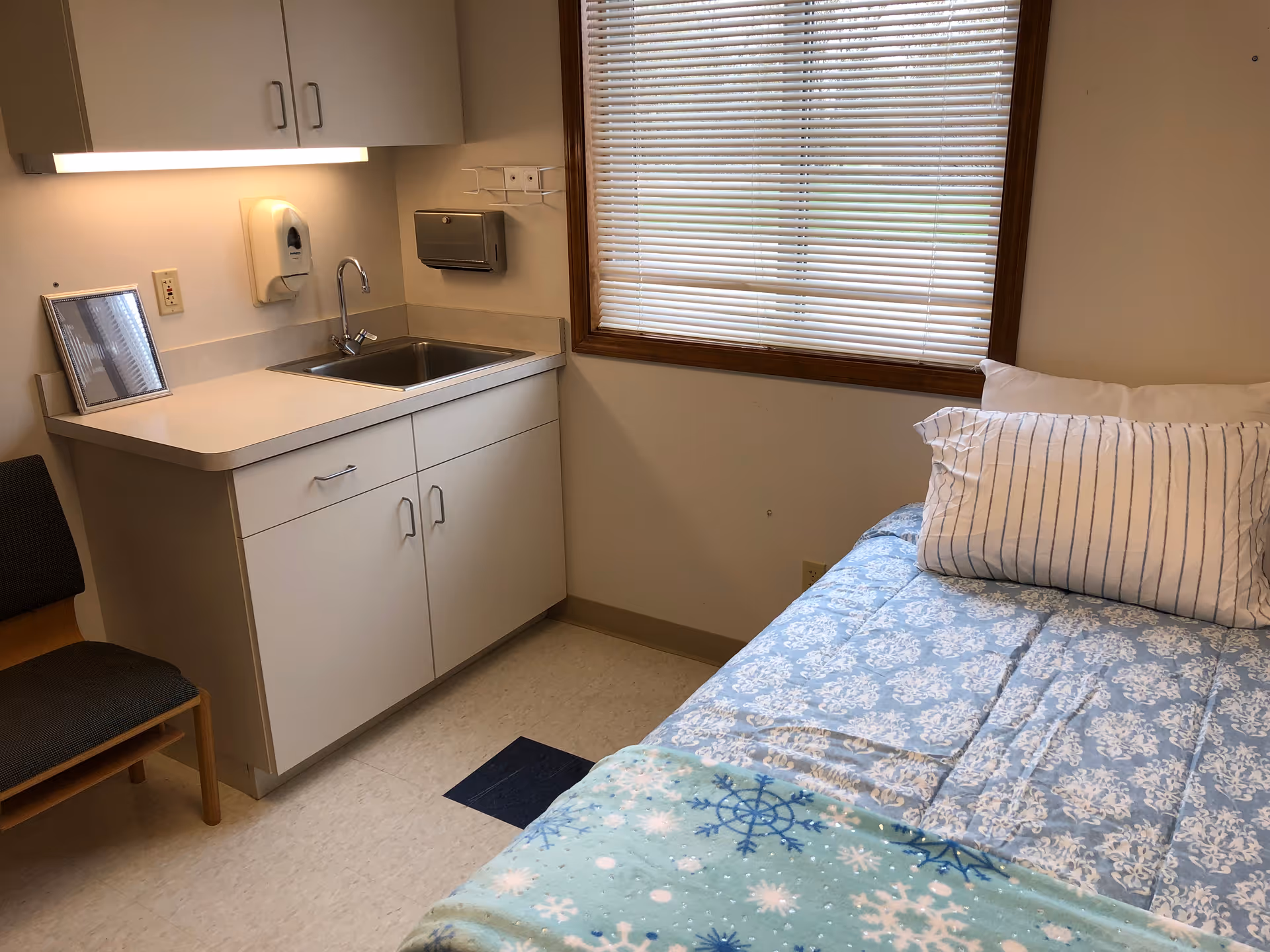 Small assisted-living bedroom with a single bed and striped pillow, a sink and cabinets under a window with blinds.