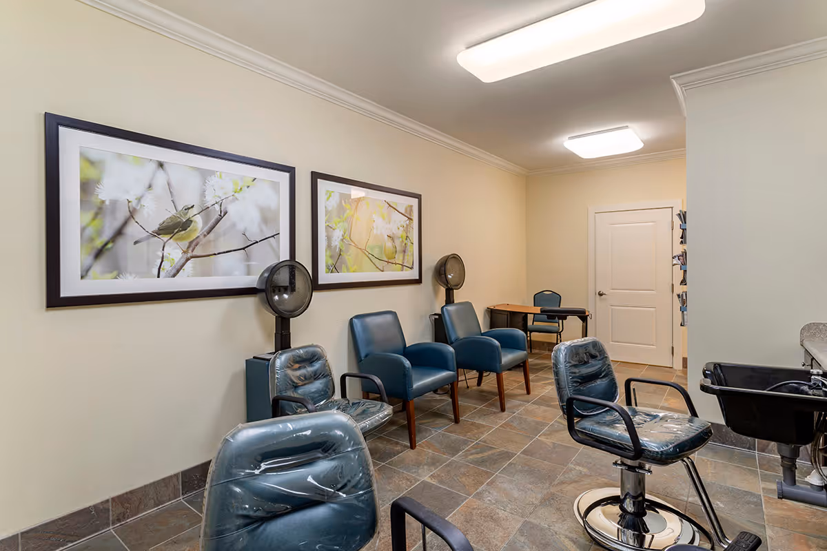 Interior view of a salon area in an assisted living facility featuring salon chairs with protective covers, hair dryers mounted on stands, blue armchairs along the wall, two framed pictures of birds on branches, a small desk and chair in the background, and a sink for hair washing.