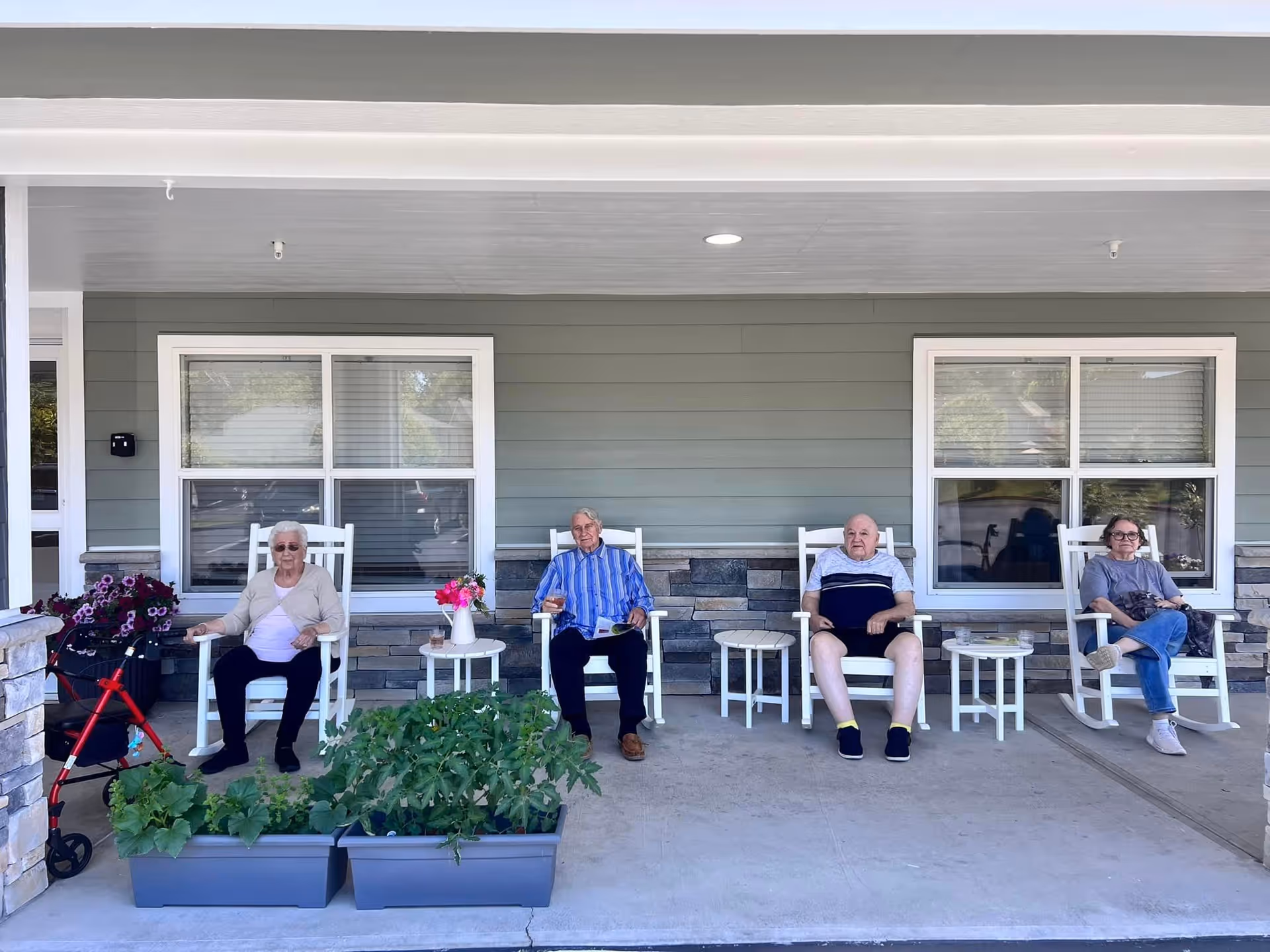 Four elderly individuals sitting on white rocking chairs on a covered porch outside a building with green siding and stone accents. There are two windows behind them, small white tables between the chairs, and two planters with green plants in front of the porch. A red walker is visible next to the person on the far left.