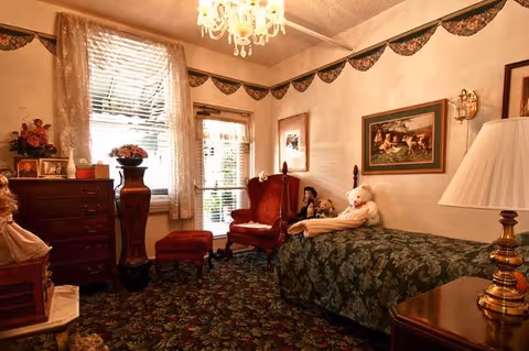 Cozy vintage bedroom with a single bed, patterned carpet and wallpaper, dresser and upholstered chairs near a window.