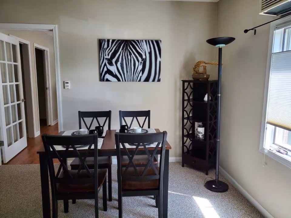 Dining room with a wooden table set for four, zebra wall art, a tall floor lamp, and a shelving unit by a window.