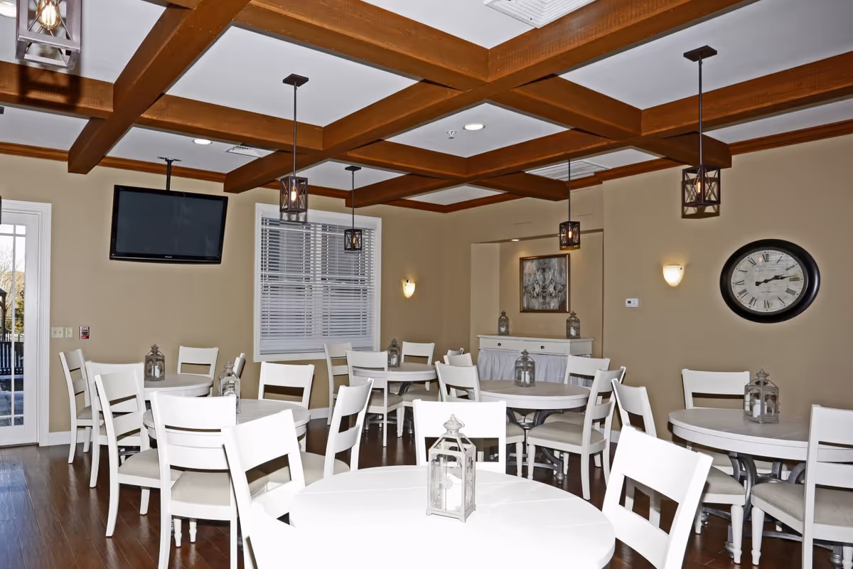 A dining room with multiple round white tables and white chairs arranged neatly. Each table has a decorative lantern centerpiece. The room features wooden ceiling beams, hanging pendant lights, a wall-mounted flat screen TV, a large clock on the wall, and a sideboard with framed artwork above it. The walls are painted beige and the floor is wooden.