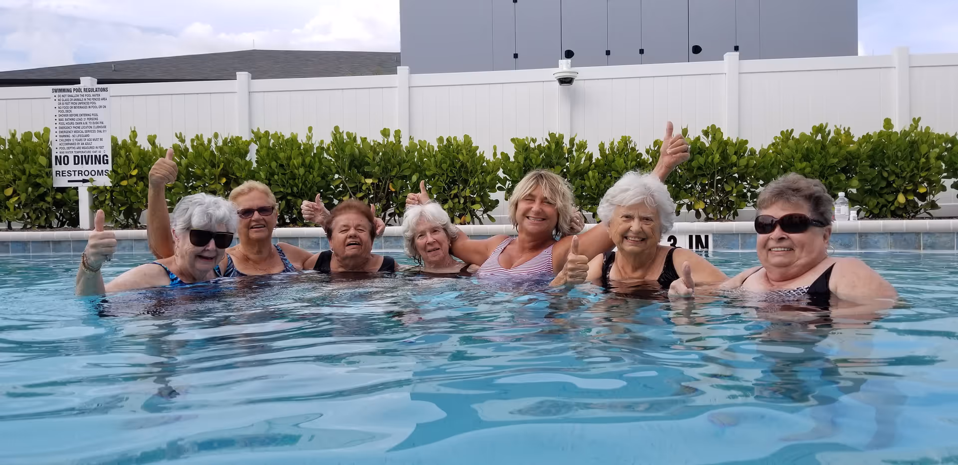 Seven elderly women in swimsuits standing in a swimming pool, smiling and giving thumbs up, with a white fence and green bushes in the background.