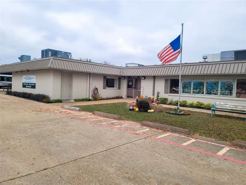 Exterior view of Gilmer Nursing & Rehabilitation facility showing a single-story building with a beige facade and a metal roof. There is an American flag on a flagpole in front of the building, along with a small garden area decorated with pumpkins and other fall-themed items. A bench is visible near the garden, and the sky is overcast.