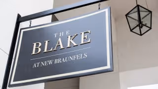 A hanging black and white sign reading "THE BLAKE AT NEW BRAUNFELS" mounted on the exterior of a building beneath a lantern-style light.