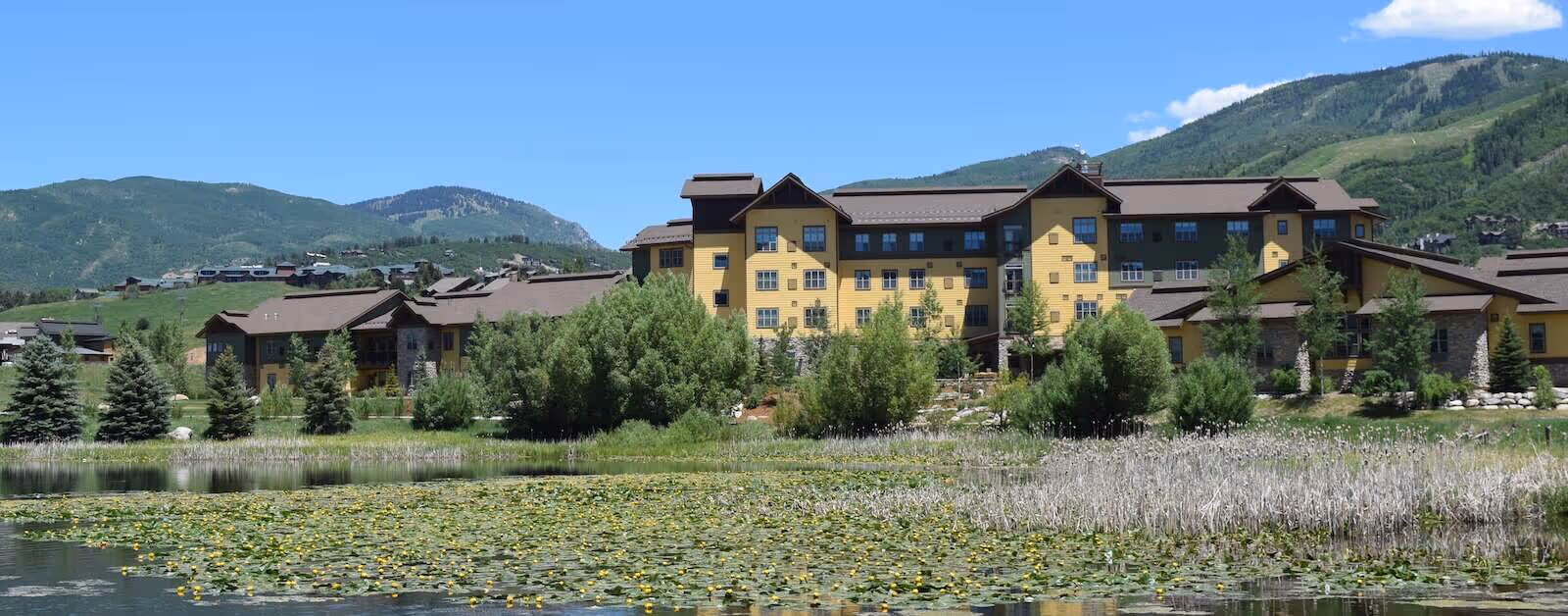 Large yellow multi-story building by a pond with lily pads, trees, and green hills in the background.