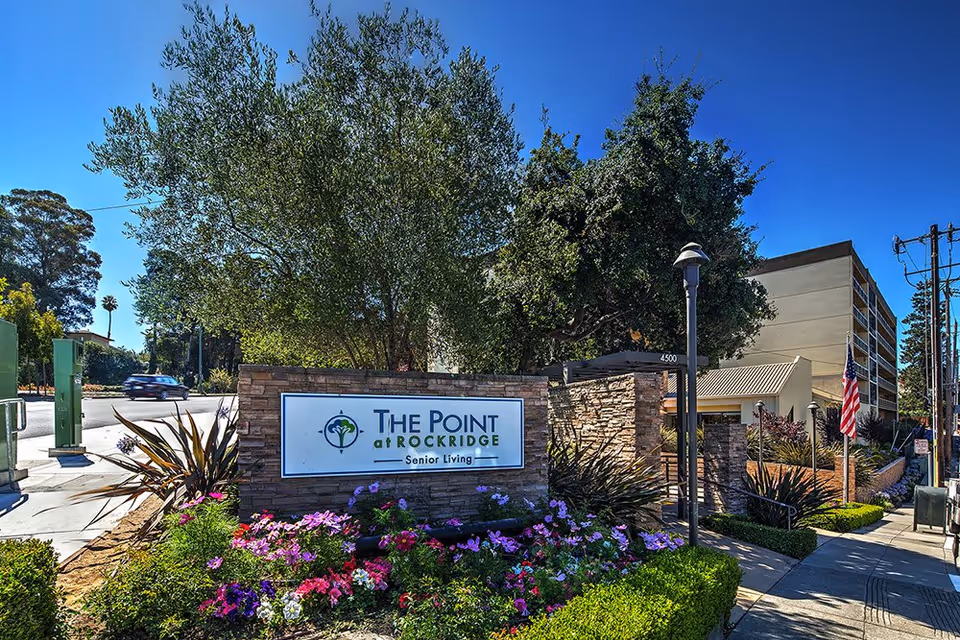Exterior view of The Point At Rockridge Senior Living facility with a stone sign displaying the facility's name surrounded by colorful flowers and greenery, a sidewalk, and a multi-story building in the background under a clear blue sky.