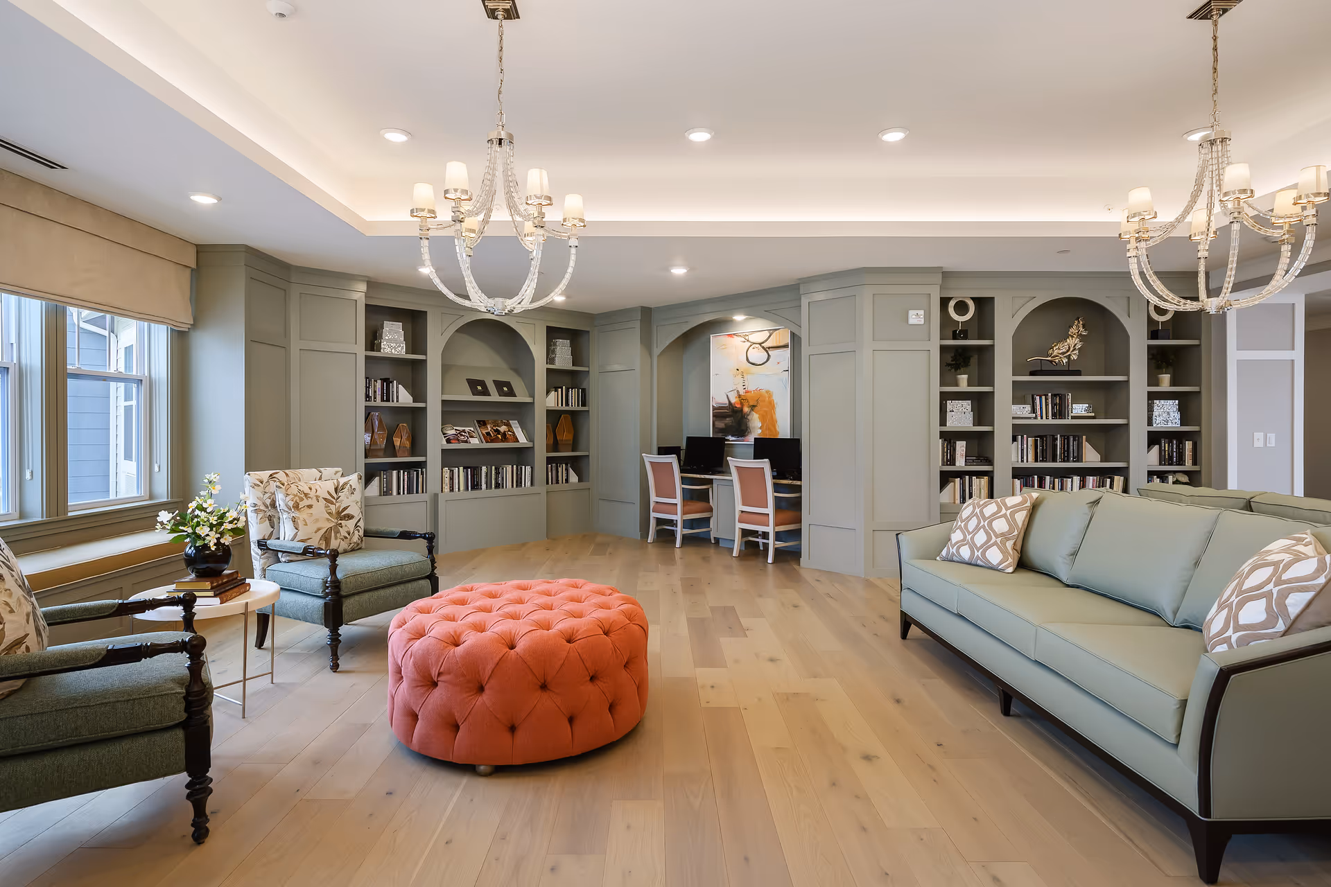 A spacious and elegant living room area with light wood flooring, a large green sofa with patterned pillows, two green armchairs with floral cushions, and a round tufted coral ottoman in the center. The back wall features built-in shelves filled with books and decorative items, and a small workspace with two chairs and computers. Two chandeliers hang from the ceiling, and large windows on the left side allow natural light to fill the room.