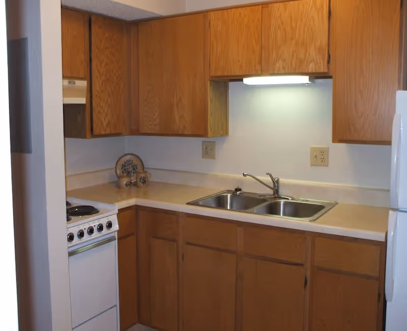 Small kitchen with wooden cabinets, a double stainless steel sink, a white stove with four burners, and a white refrigerator. There is a decorative plate and small items on the countertop in the corner.