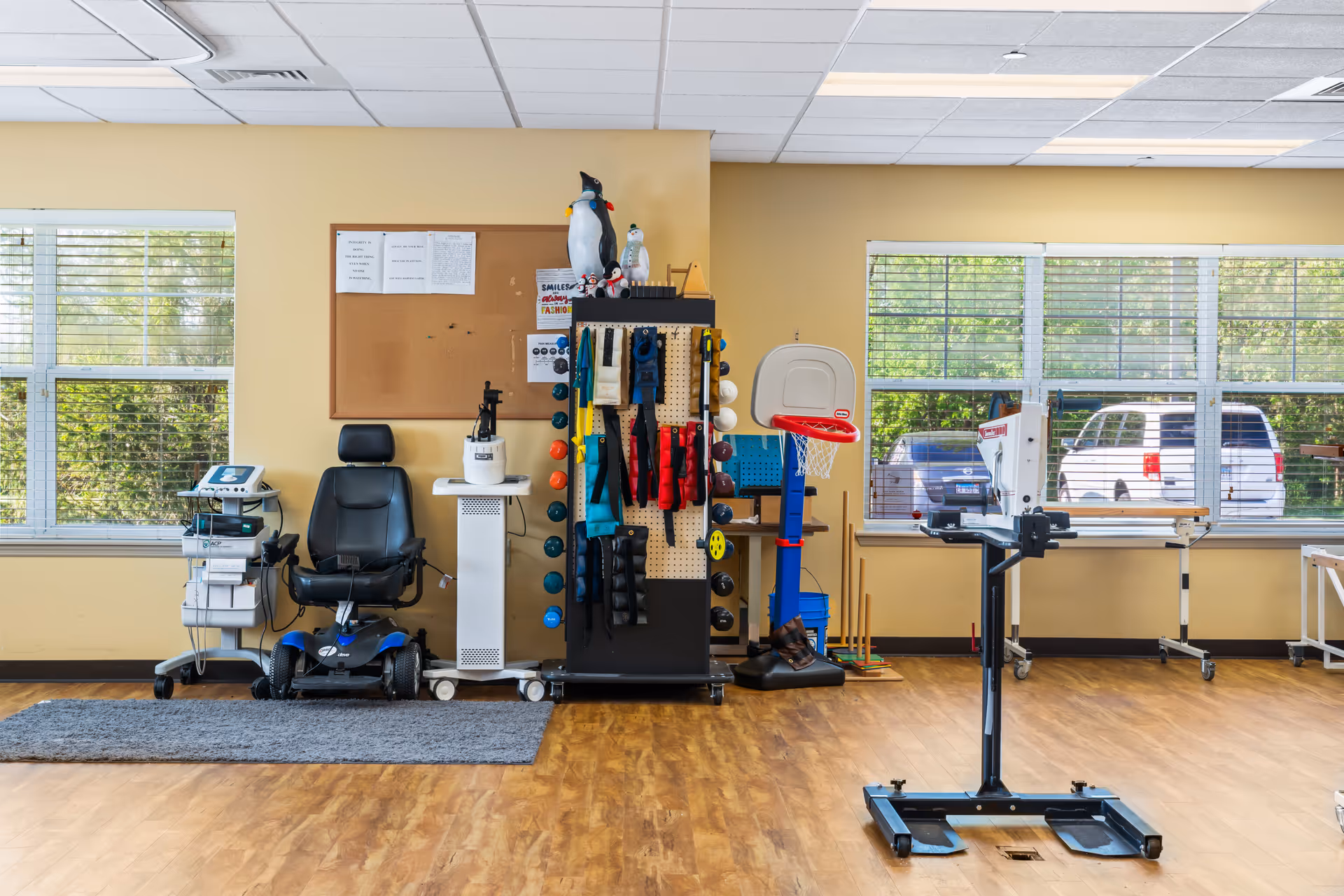 A rehabilitation or therapy room with exercise and medical equipment including a mobility scooter, therapy tools on a rack, a basketball hoop, and various medical devices. The room has large windows with blinds, yellow walls, and a wooden floor.