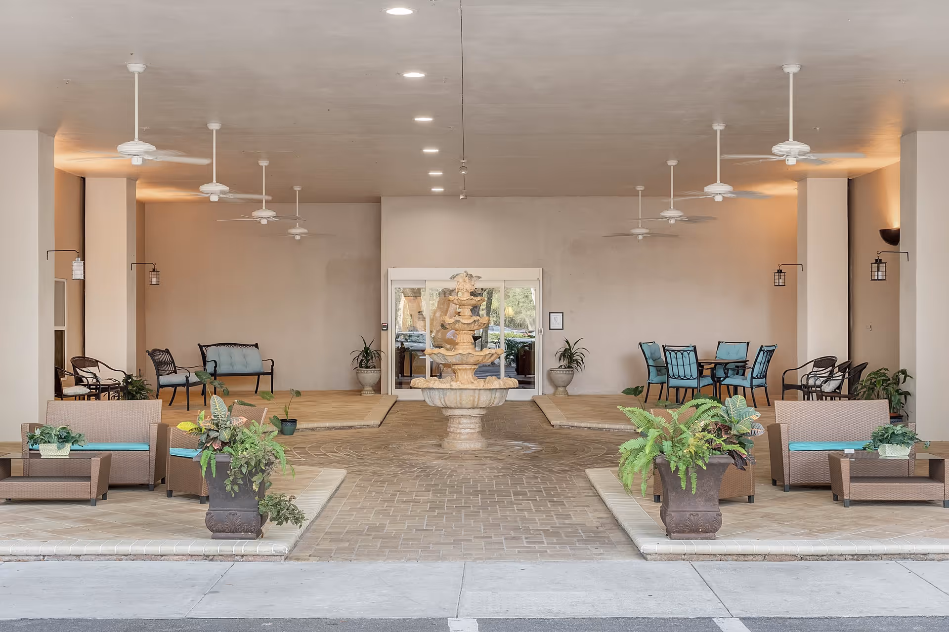 Covered outdoor seating area with wicker furniture, potted plants, ceiling fans, and a central stone fountain. There are multiple seating arrangements including cushioned chairs and tables with chairs, set against a beige wall with soft lighting.