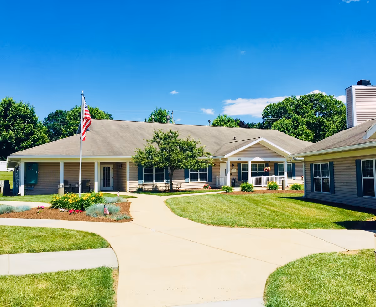 Exterior view of a single-story senior living facility building with beige siding, a gray roof, and several windows. There is a concrete walkway leading to the entrance, a flagpole with an American flag, green grass, and landscaped flower beds under a clear blue sky.