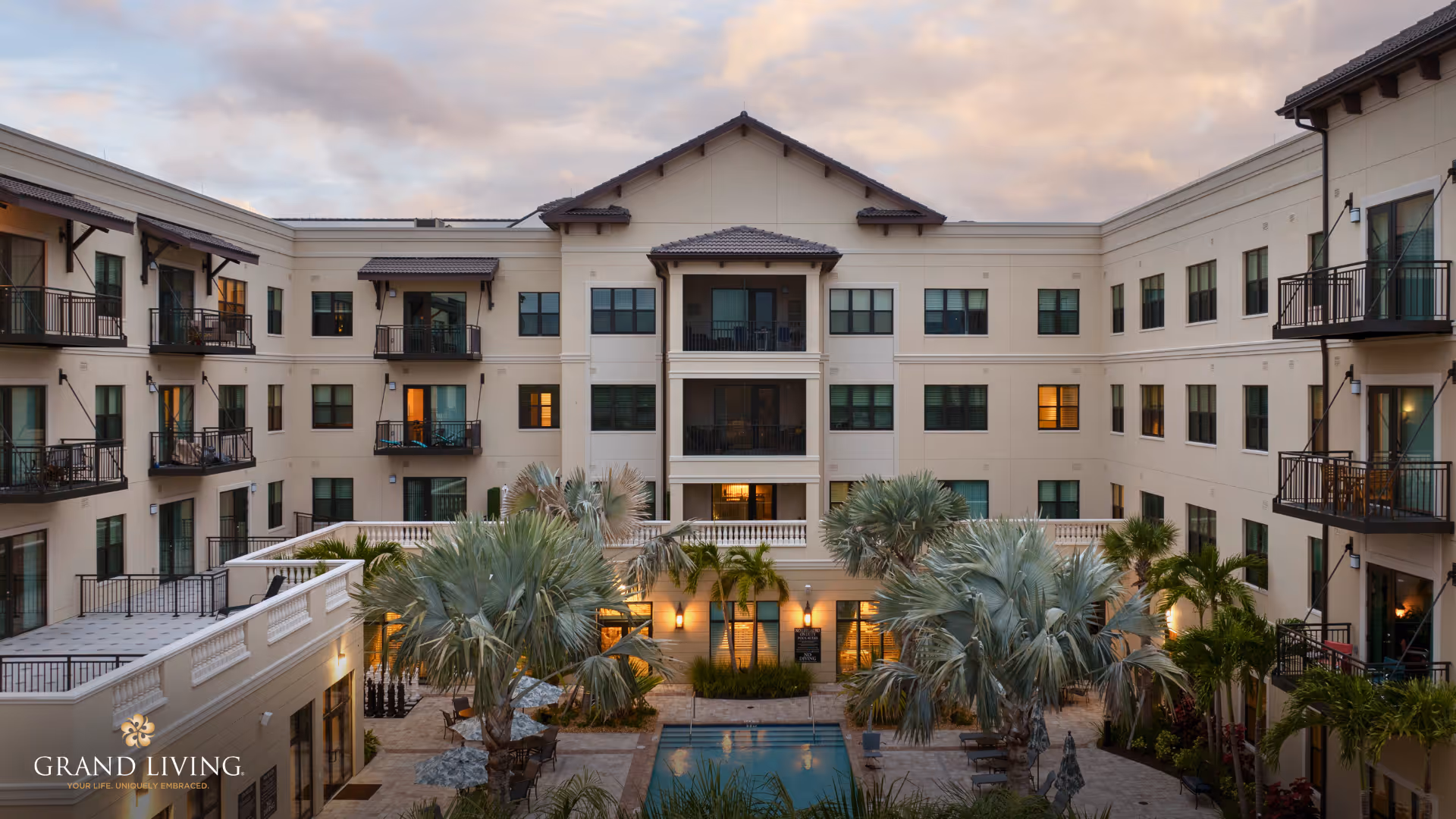 View of the courtyard of a senior living facility named Grand Living at Wellen Park, featuring a central swimming pool surrounded by palm trees and lounge chairs, with a multi-story building with balconies and windows enclosing the courtyard.