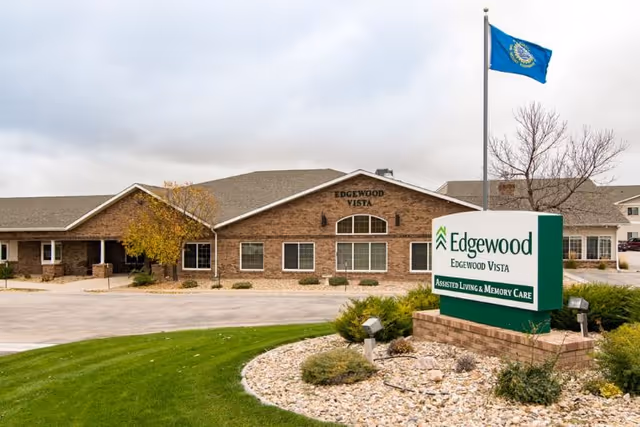 Exterior view of Edgewood Vista, a single-story brick building with a sign in front that reads 'Edgewood Edgewood Vista Assisted Living & Memory Care.' There is a flagpole with a blue flag and landscaped greenery and rocks around the sign.