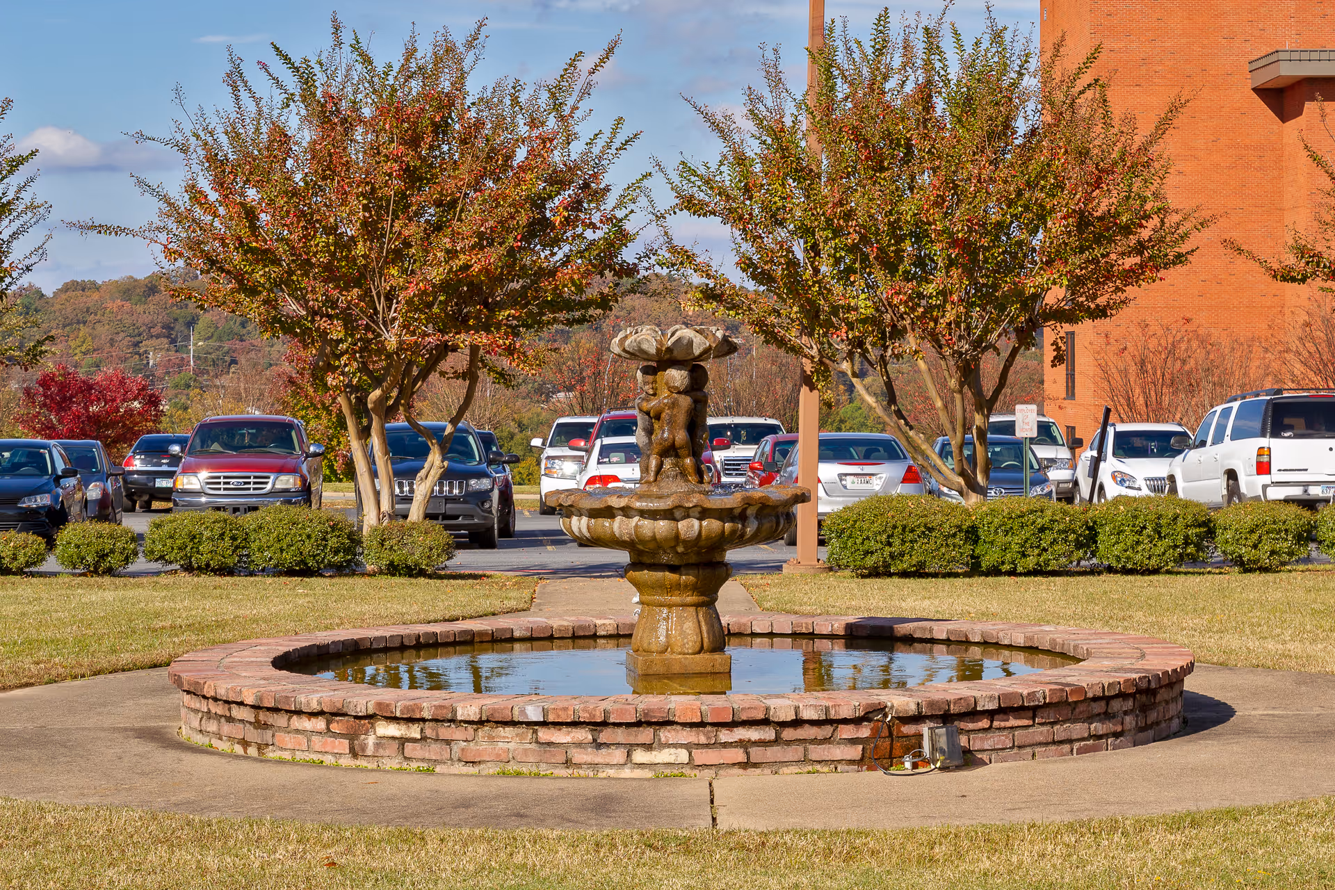 A circular brick water fountain with a sculpted centerpiece featuring cherubs, situated in a grassy area with trimmed bushes and trees with autumn foliage. Behind the fountain is a parking lot with several parked cars and a large brick building on the right side under a partly cloudy sky.