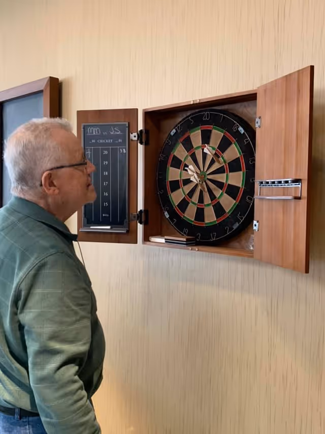 An elderly man wearing glasses and a green sweater stands indoors looking at a dartboard mounted on a wall. The dartboard is inside a wooden cabinet with open doors, and several darts are stuck in the board. Next to the dartboard is a scoreboard for the game of cricket.