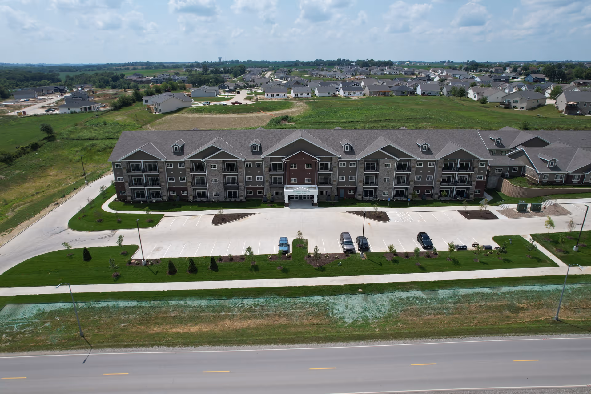 Aerial view of a large three-story residential building with multiple balconies and a parking lot in front. The building is surrounded by green lawns and is located in a suburban area with many houses and open fields in the background under a partly cloudy sky.