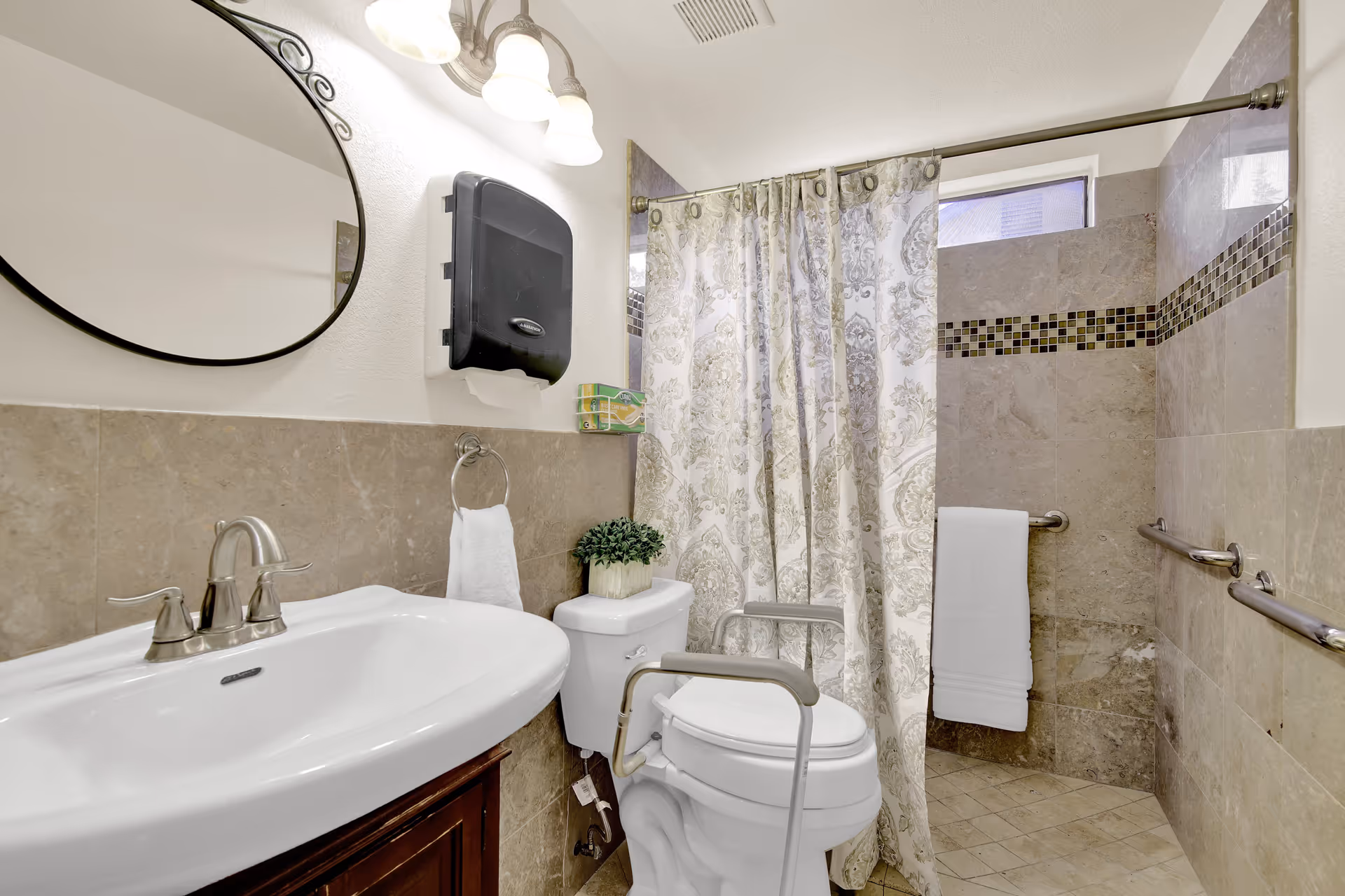 A bathroom with beige tiled walls and floor, featuring a white sink with silver faucet, an oval mirror above the sink, a toilet with safety rails, a decorative plant on the toilet tank, a patterned shower curtain, and grab bars on the walls near the shower area.