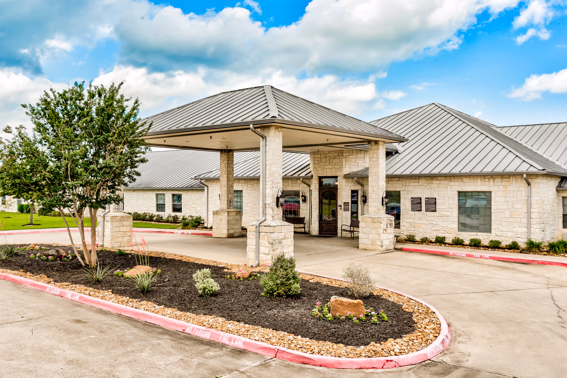 Stone-faced nursing facility entrance with a covered porte-cochere, landscaped planting island, and metal roof under a partly cloudy sky.