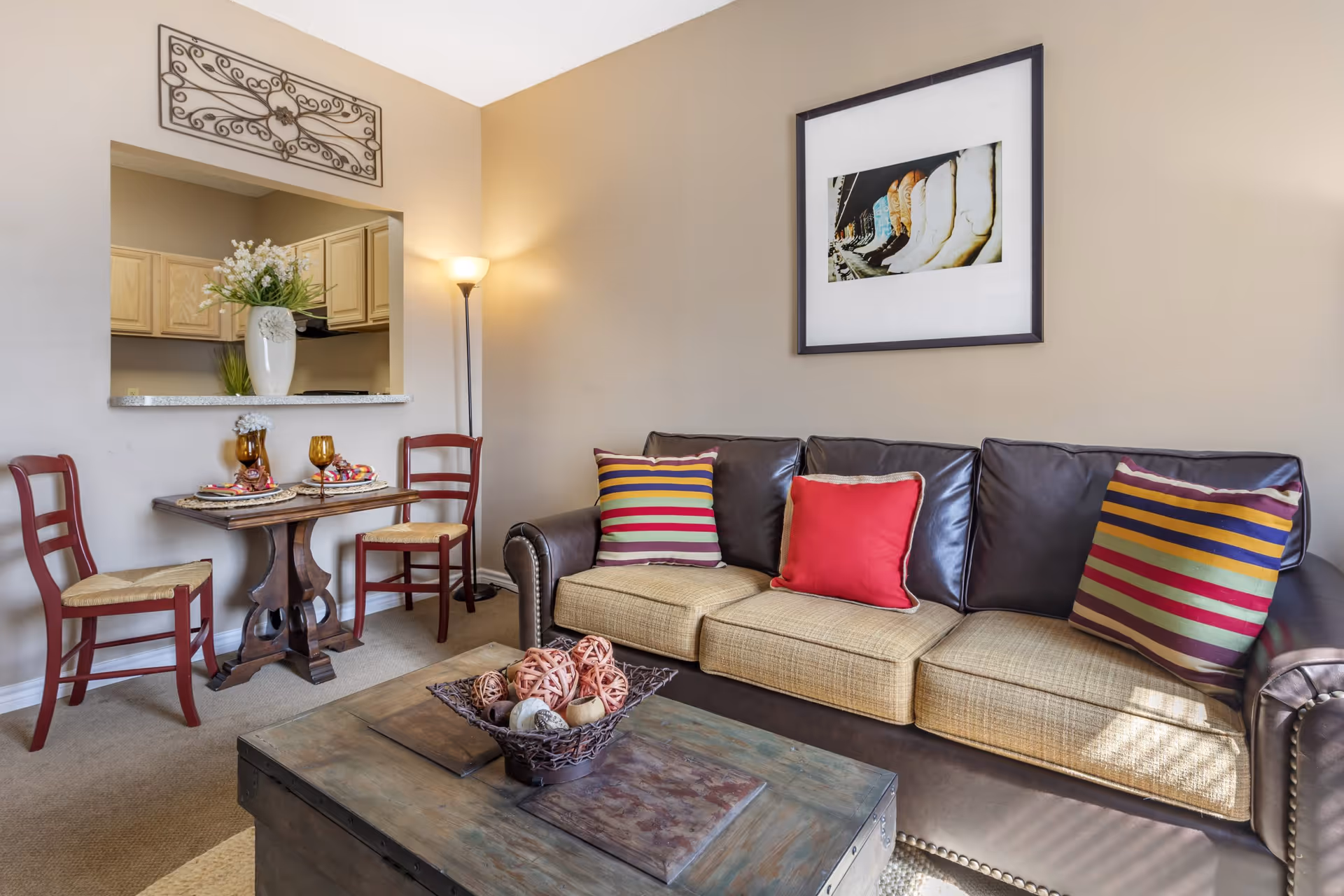 Cozy living room with a leather-and-fabric sofa topped with colorful pillows, a wooden coffee table centerpiece, and a small dining nook with two red chairs and a kitchen pass-through.