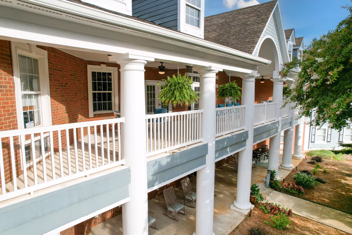 Exterior view of a senior living facility with a two-story porch featuring white columns and railings. The upper porch has hanging green plants and seating, while the lower porch has rocking chairs. The building has red brick walls and white-framed windows, with a landscaped area including bushes and flowers nearby.