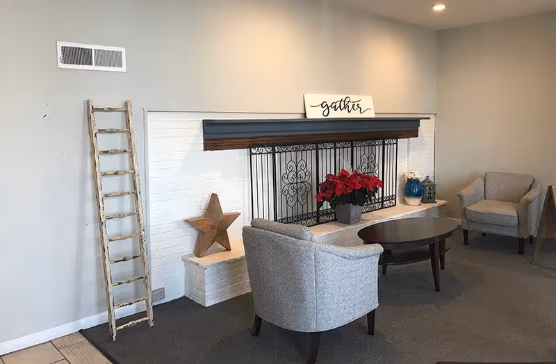 A cozy living room area with two gray upholstered armchairs and a dark wooden coffee table in front of a white brick fireplace. The fireplace has a decorative black metal screen and a wooden mantel with a sign that reads 'gather'. There is a wooden star decoration on the left side of the fireplace and a potted red poinsettia plant on the hearth. A rustic wooden ladder leans against the wall to the left of the fireplace. The walls are painted light gray and the floor is carpeted.