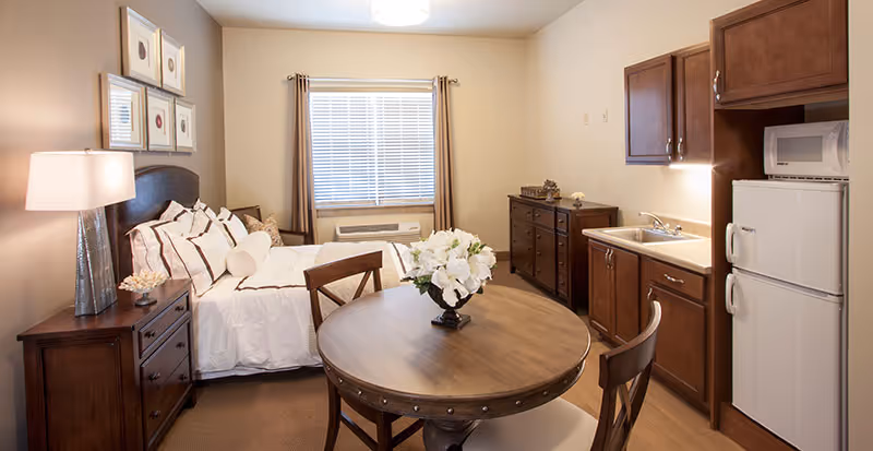 Studio apartment interior with a bed, round dining table, and a kitchenette with fridge and cabinets.