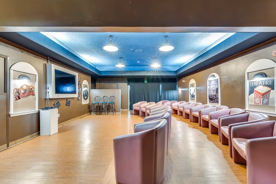 Interior view of a theater room in a senior living facility with rows of pink armchairs facing a large flat-screen TV mounted on a brown wall. The room has a blue ceiling with recessed lighting and movie-themed decorations on the walls, including posters and a popcorn illustration labeled 'THEATRE'.