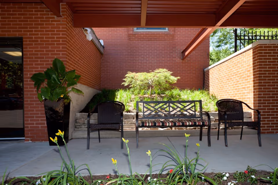 Covered outdoor seating area with a bench and two chairs in front of a brick wall and planted landscaping.