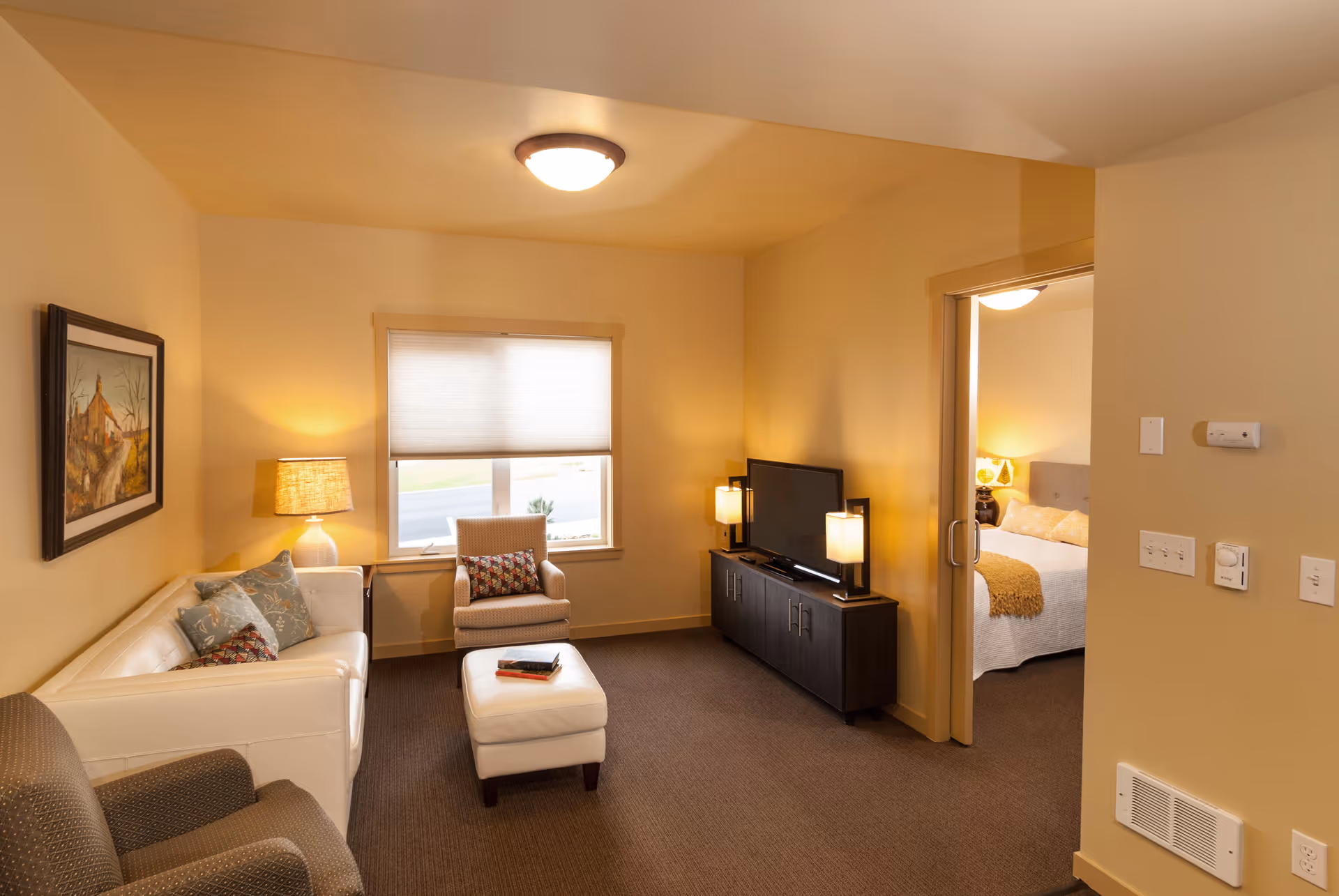 A cozy living room area with a white couch adorned with decorative pillows, a beige armchair, and a matching ottoman. A window with a closed blind is centered on the back wall. A dark wood TV stand holds a flat-screen television flanked by two lamps. To the right, an open doorway leads to a bedroom with a bed, nightstand, and lamp visible. The walls are painted a warm beige color.