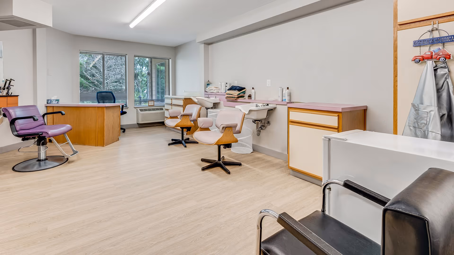 Interior of a hair salon area in a senior living facility with multiple salon chairs, sinks for washing hair, a wooden desk with an office chair near large windows, and hair care products on the counter.