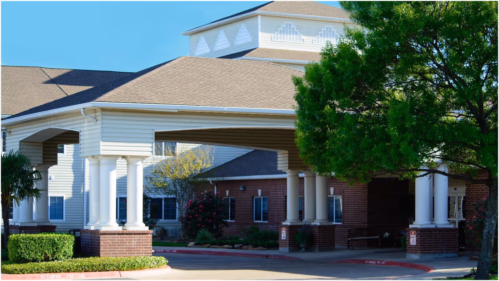 Exterior view of The Court at Round Rock senior living facility showing a covered entrance with white columns, brick and siding walls, and surrounding greenery including trees and bushes under a clear blue sky.