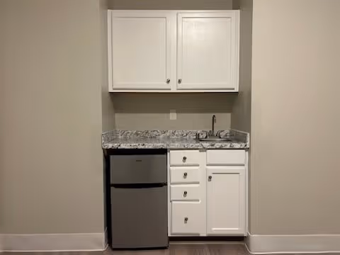 Small kitchenette area with white cabinets, a granite countertop, a stainless steel mini refrigerator, and a small sink with a faucet, set against a beige wall.